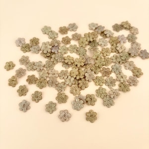 Peut inclure: Une collection de petites fleurs en crochet, de différentes nuances de vert, de gris et d'argent. Les fleurs sont dispersées sur une surface beige clair, mettant en valeur leurs motifs délicats et détaillés. Idéal pour les projets d'artisanat.