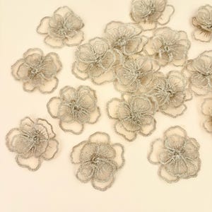 May include: A collection of silver-toned floral appliques featuring delicate, sheer mesh petals with metallic thread embroidery. Each flower has a layered, scalloped edge design with a textured center, arranged in a scattered pattern.