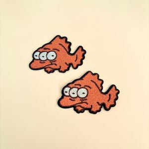 Op de afbeelding: Twee oranje geborduurde vispatches met elk drie ogen. De patches hebben een zwarte rand en zijn geplaatst tegen een cr&egrave;mekleurige achtergrond. De vissen hebben een cartoonachtig ontwerp.