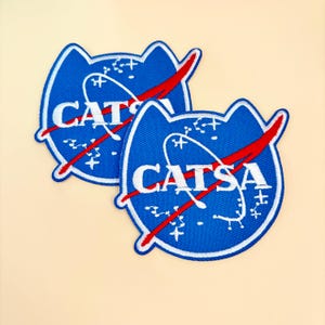 May include: Two blue embroidered patches with a cat-shaped outline. The patches feature the text "CATSA" in white, with a red swoosh and a constellation design. The patches are on a light beige background.
