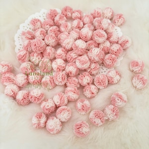 Puede incluir: Una colección de pequeños pompones rosas y blancos, esparcidos sobre un mantel individual blanco y calado y una superficie blanca y esponjosa. Los pompones parecen estar hechos de un material suave y texturizado. La imagen está bien iluminada.