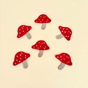 May include: Six embroidered mushroom patches with red caps and white spots, each with a gray stem. The patches are arranged on a cream-colored background. These patches are perfect for adding a whimsical touch to clothing or accessories.
