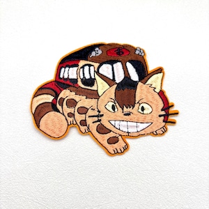 May include: An embroidered patch featuring a tan and brown cat-like creature with a wide, toothy grin and large yellow eyes. The creature has a long, striped tail and is attached to a brown, bus-shaped body with white windows and a red roof.