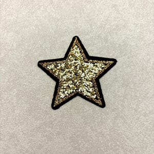 May include: A gold glitter star patch with a black border. The star is filled with gold glitter and has a black outline. The patch is on a light gray background. This patch can be used to decorate clothing or accessories.