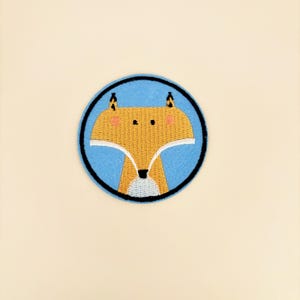 May include: A circular embroidered patch featuring a stylized fox head with golden-yellow fur, black-tipped ears, pink cheeks, and a white chest area, all set against a light blue background enclosed by a thick black circular border.