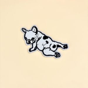 May include: Embroidered patch featuring a stylized French Bulldog in a reclining pose. The dog is primarily white with black spots and a black outline. The patch is on a light beige background.