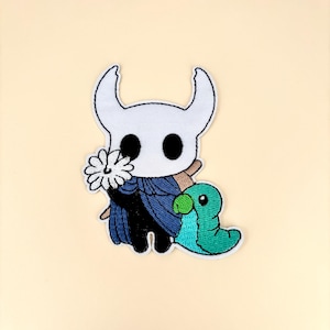 May include: Embroidered patch featuring a white character with large horns, black eyes, and a blue cape, holding a white flower and accompanied by a green creature. The patch is on a cream-colored background.