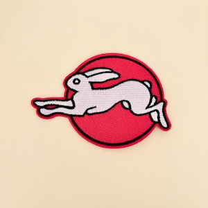 May include: A circular embroidered patch featuring a white rabbit in a leaping position against a solid red background. The rabbit is outlined in black thread, and the entire circular design is bordered by a thick black stitched edge.