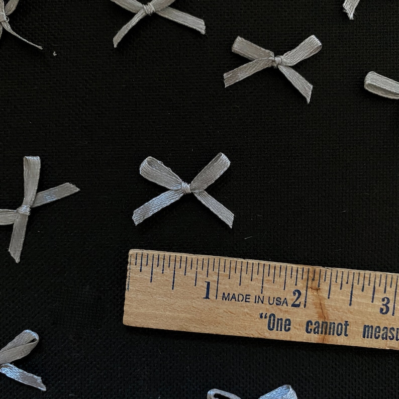 Puede incluir: Primer plano de una regla que mide 7,6 cm con el texto "Made in USA" y "One cannot measure" impreso en ella. La regla est&aacute; sobre una superficie negra con varios lazos de sat&eacute;n plateado esparcidos a su alrededor.