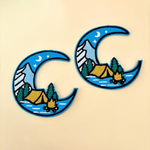 May include: Two blue crescent moon-shaped embroidered patches with a camping scene inside. The scene includes a tent, a campfire, trees, and mountains. The moon has a white crescent moon and a star inside.