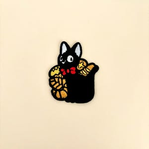 May include: Embroidered patch featuring a black cat with white ears and eyes, a red bow tie, and holding a bundle of gold and yellow objects. The patch is on a cream-colored background.