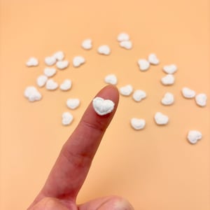 May include: A close-up shot of a finger holding a small, white, heart-shaped embellishment. Scattered around are several other similar heart-shaped decorations. The background is a solid, light orange color. These could be used for crafts or decorations.