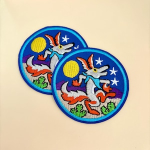 May include: Two embroidered iron-on patches featuring a coyote howling at the moon in a desert landscape. The coyote is white with orange and red accents. The moon is yellow and the sky is blue with white stars. The patches are circular and have a blue border.