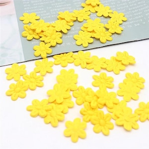 May include: A collection of small, bright yellow daisy-shaped embellishments scattered on a white surface. The flowers have detailed petals and a textured center. Some are on a printed paper with text.