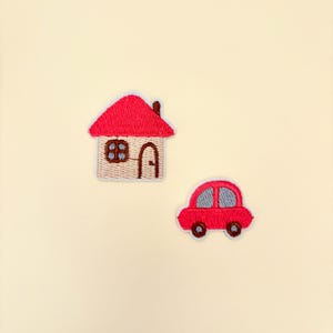 May include: Embroidered patches featuring a red house with a brown chimney and a red car with gray windows. The house has a tan body with a brown door and window. The car has black wheels and gray windows. The patches are on a cream background.