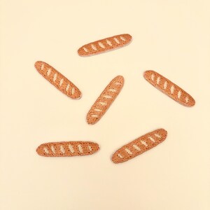 May include: A set of six embroidered patches shaped like small baguettes. Each patch features a golden-brown thread colour with lighter cream-coloured diagonal slashes representing the crust texture of the bread loaves in a long, thin shape.