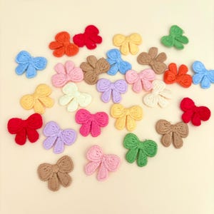 May include: A collection of colorful, embroidered bow appliques in various shades, including red, pink, blue, yellow, and green. These decorative bows are arranged on a light beige surface, perfect for crafting and embellishments.