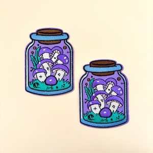 May include: Two embroidered iron-on patches of purple mushrooms in glass jars with a light purple background.