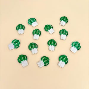 May include: A collection of embroidered patches shaped like bok choy. Each patch features a green leafy top and a white base. The patches are scattered on a light beige background.