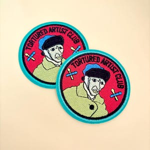 May include: Two round embroidered patches with a red background and a teal border. The patches feature a cartoon image of a person wearing a blue hat and green coat. The text "TORTURED ARTIST CLUB" is in a circle around the image.