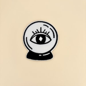 May include: A white embroidered patch featuring a crystal ball design with an eye in the center. The eye has a diamond-shaped pupil and is surrounded by eyelashes. The patch has a black border and base, set against a cream background.