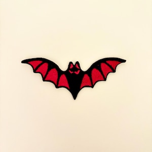 May include: Embroidered bat patch in black with red wing details. The bat has a stylized design with a pointed head and spread wings. This patch is suitable for Halloween or gothic-themed projects.
