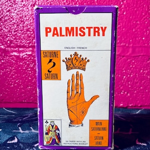 Puede incluir: Una caja morada con las palabras "Palmistry" en letras naranjas. La caja contiene 56 cartas con un folleto de instrucciones. La parte delantera de la caja presenta una mano con líneas y símbolos, una corona con el número 5, y las palabras "Saturne" y "Saturn".