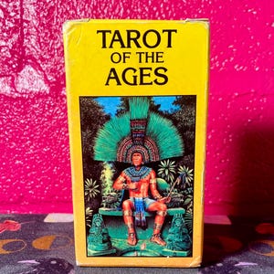 Puede incluir: Una caja amarilla con el texto "Tarot of the Ages" en la parte delantera. La caja presenta una ilustración de una persona con un tocado verde sentada en un trono.