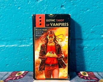 May include: A black box with a red and yellow background image of a person with long dark hair holding a small green object. The box has the text "Gothic Tarot of Vampires" in white letters. The text "Riccardo Minetti" and "Artwork by Emiliano Mammucari" is also on the box.