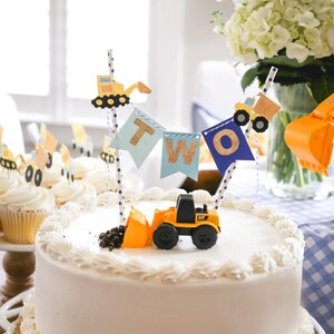May include: A white cake decorated with blue and yellow construction vehicle themed decorations. The cake is topped with a banner that says "TWO" and a small toy construction vehicle.