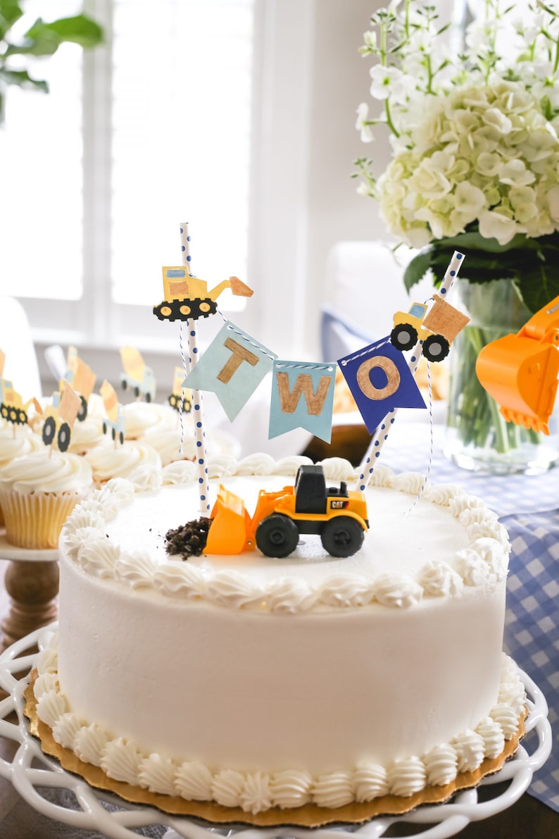 May include: A white cake decorated with blue and yellow construction vehicle themed decorations. The cake is topped with a banner that says "TWO" and a small toy construction vehicle.