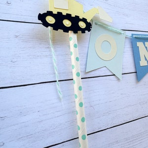 May include: A yellow construction vehicle with black wheels and a blue and white window is attached to a string of blue and white flags that spell out "ONE". The string is attached to a white straw with green polka dots.