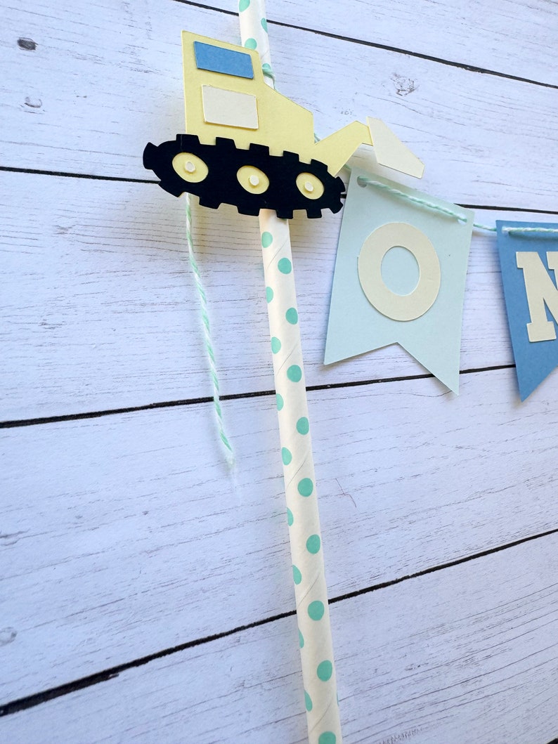 May include: A yellow construction vehicle with black wheels and a blue and white window is attached to a string of blue and white flags that spell out "ONE". The string is attached to a white straw with green polka dots.
