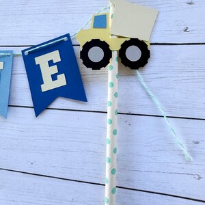 May include: A blue and yellow paper banner with the letter "E" on one of the flags. A yellow toy truck with a white bed is attached to a white and green striped straw. The straw is attached to the banner with white string.
