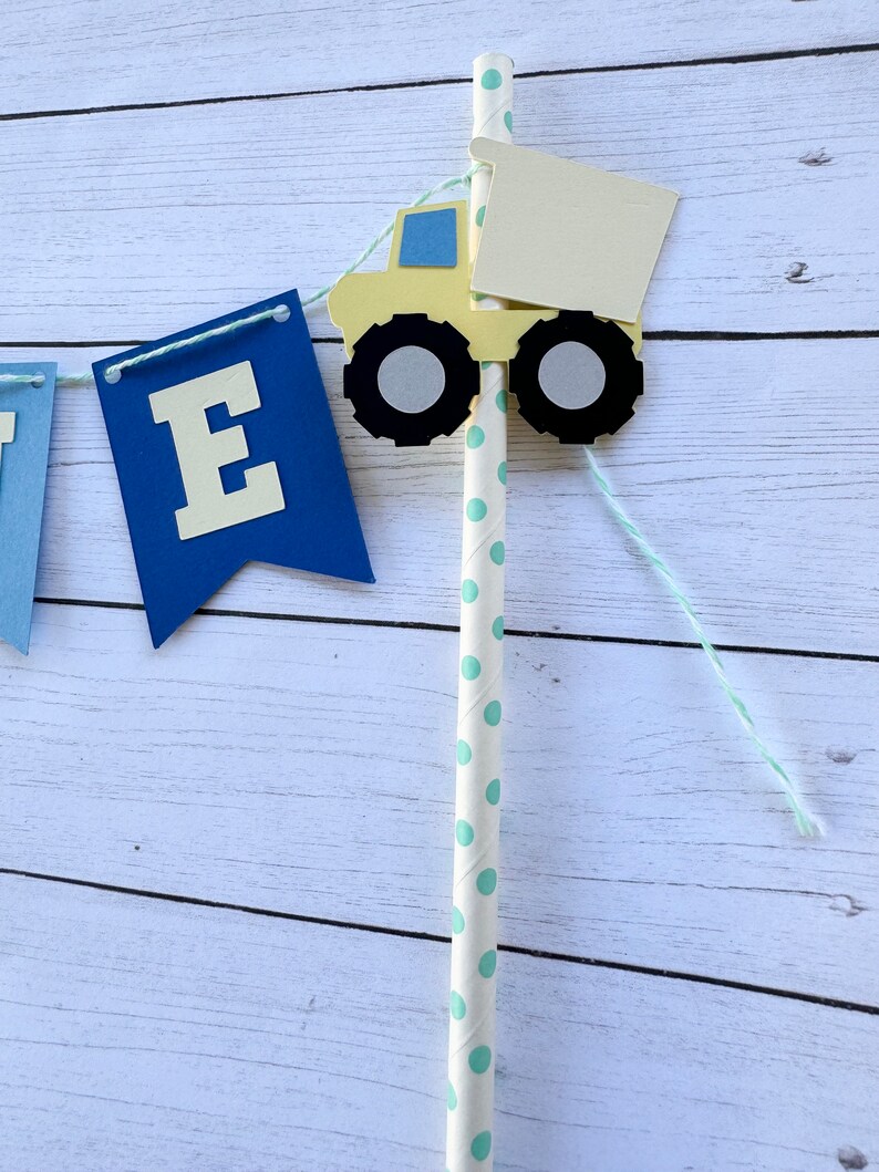 May include: A blue and yellow paper banner with the letter "E" on one of the flags. A yellow toy truck with a white bed is attached to a white and green striped straw. The straw is attached to the banner with white string.