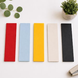 May include: Five rectangular bookmarks arranged in a row, featuring solid colors of red, light blue, yellow, cream, and black. Each bookmark has a smooth, flat surface and straight edges, presented in a clean, organized layout for easy viewing.