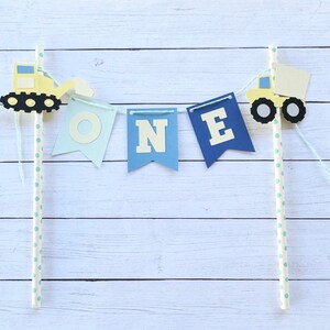 May include: A blue and yellow construction-themed cake topper with the word "ONE" on flags and two toy trucks. The flags are attached to white straws with blue polka dots.