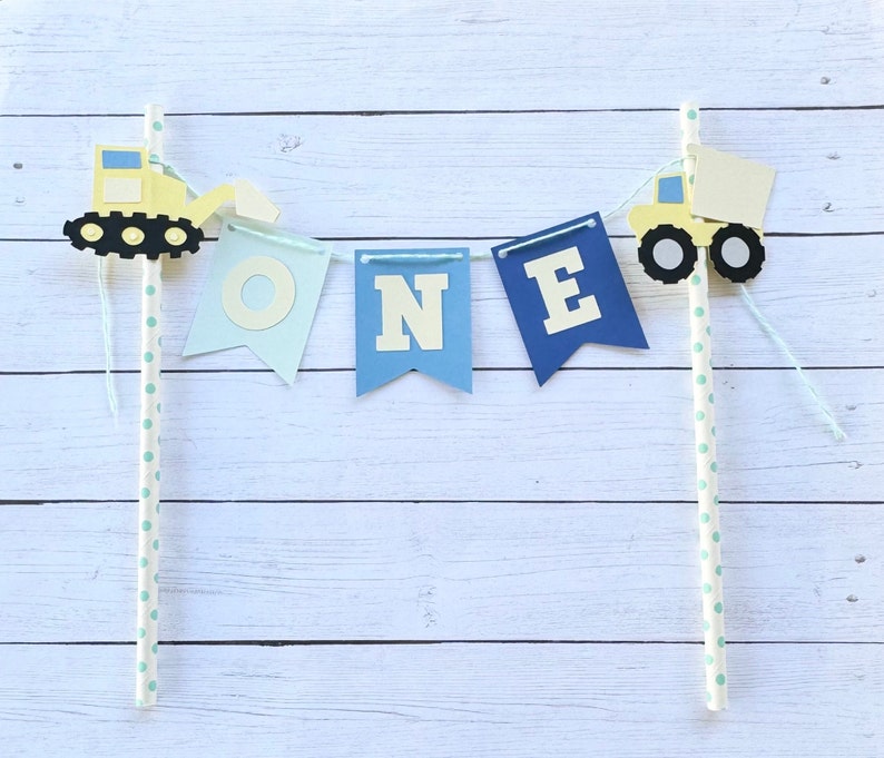 May include: A blue and yellow construction-themed cake topper with the word "ONE" on flags and two toy trucks. The flags are attached to white straws with blue polka dots.