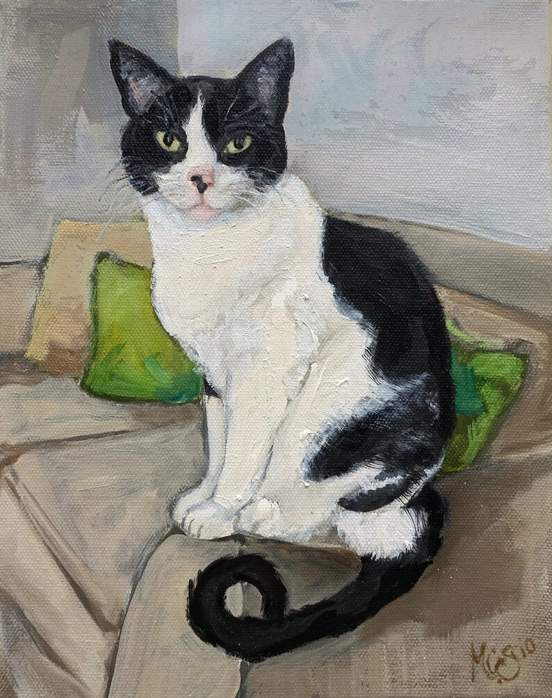 May include: A black and white cat with green eyes sits on a brown couch with green pillows. The cat's tail is curled around its body.