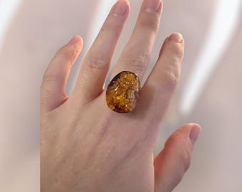 May include: A large, oval-shaped amber ring. The ring features a translucent, golden-brown amber stone with visible inclusions. The amber is set on a simple band, creating a statement piece. The ring is a unique piece of jewelry.