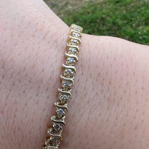 Puede incluir: Un brazalete dorado con una serie de pequeñas piedras preciosas transparentes en un patrón repetitivo. El brazalete está formado por elementos en forma de onda. El diseño es flexible y está diseñado para usarse en la muñeca. Las piedras están engastadas en oro.