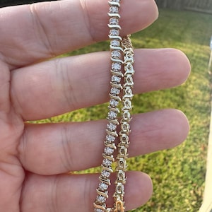 Puede incluir: Dos pulseras doradas con una serie de pequeñas piedras brillantes. Las pulseras se sostienen sobre un fondo de hierba verde y una valla borrosa. Las pulseras están conectadas con un cierre.