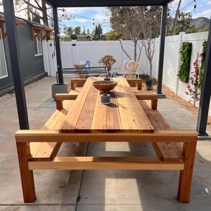 Może przedstawiać: Drewniany stół piknikowy z dołączonymi ławkami stoi pod czarną pergolą. Stół wykonany jest z jasnobrązowego drewna i ma naturalne wykończenie. Drewniana miska i dwa szare cylindry znajdują się na stole. Otoczenie jest na zewnątrz, z białym płotem i zielenią w tle.