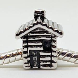 May include: Silver charm in the shape of a small log cabin with a chimney. The charm is on a silver bracelet.