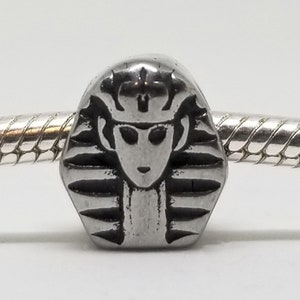 May include: A silver charm bead with a detailed depiction of an Egyptian pharaoh's head, wearing a traditional headdress.