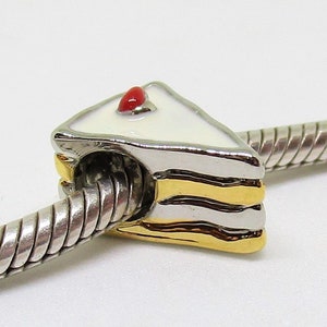 Op de afbeelding: Een witte en gouden bedel in de vorm van een stuk taart met een rode kers erop. De bedel zit op een zilveren slangenkettingarmband.