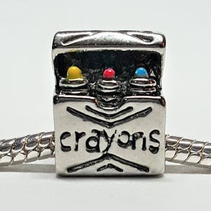 May include: A silver charm with a box design, featuring three colorful crayon tops in yellow, red, and blue. The word "crayons" is inscribed on the front. The charm is attached to a silver chain.