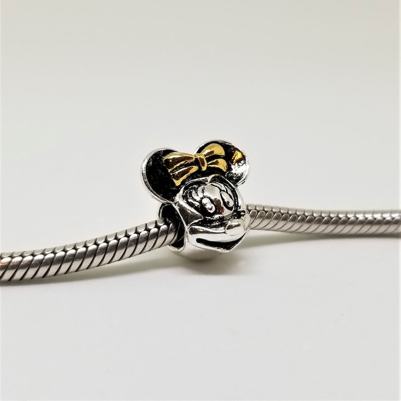 May include: Silver charm featuring a black and white image of Minnie Mouse with a gold bow.