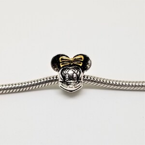 May include: Silver charm featuring a gold Minnie Mouse head, with a yellow bow and black ears.