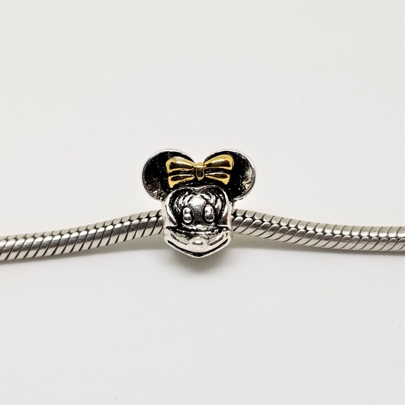 May include: Silver charm featuring a gold Minnie Mouse head, with a yellow bow and black ears.
